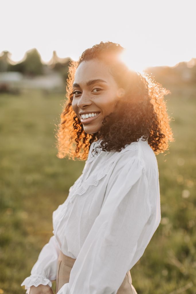 Portrait of a smiling woman with curly hair in a sunlit outdoor field.