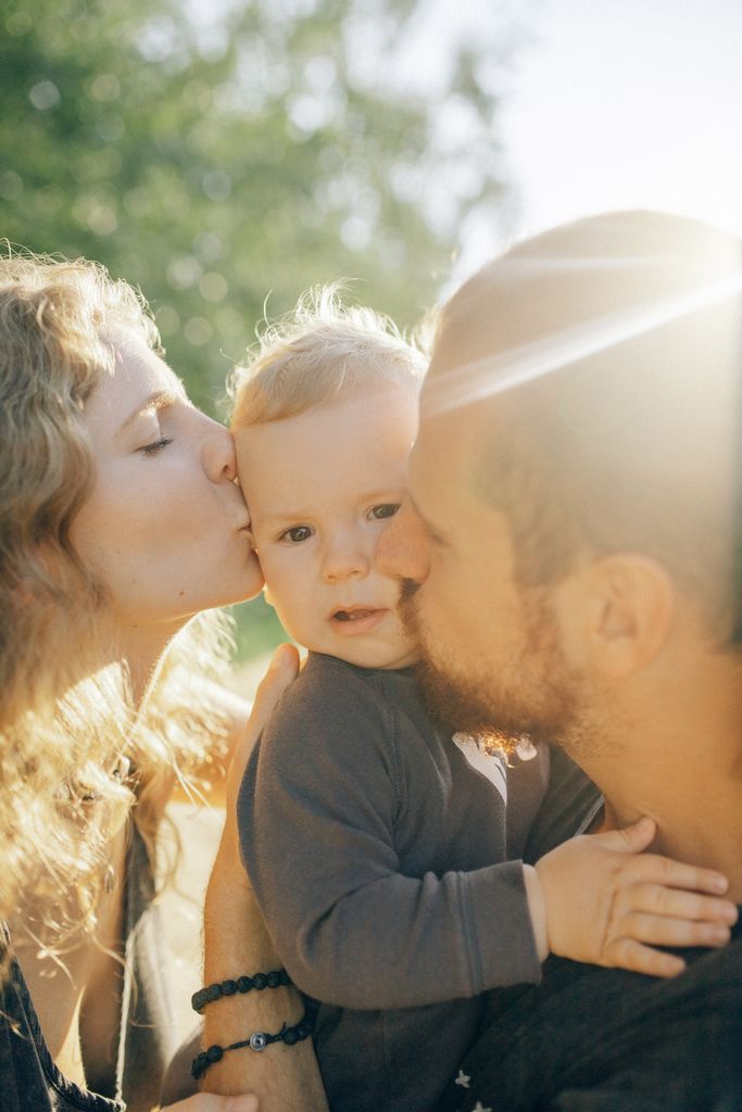 Tender family moment outdoors with parents kissing their baby, bathed in sunlight.