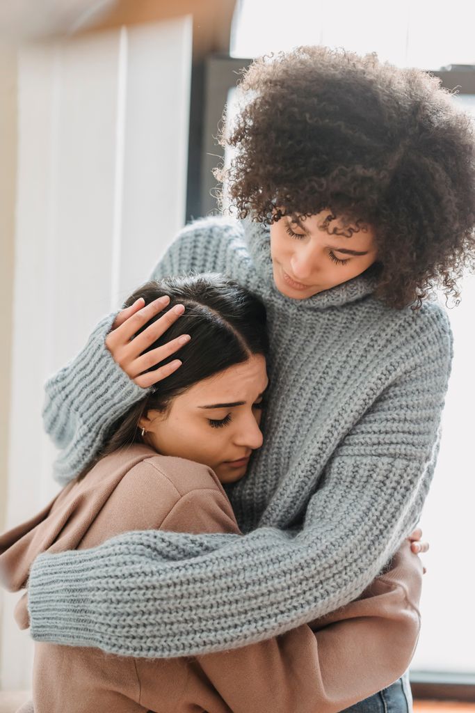 Young multiethnic female friends in casual clothes embracing each other while crying in light apartment